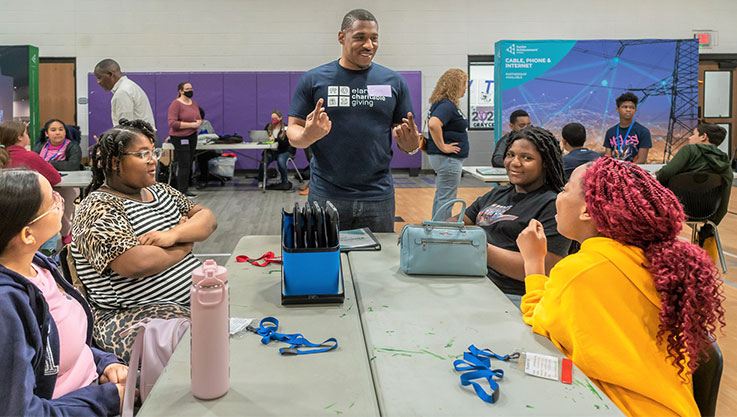 Students sit at a table and face an adult volunteer speaking to them. 