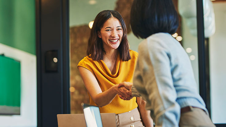 Two women stand across a conference table and share a handshake. 